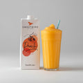 Sweetbird peach smoothie mix carton with a glass of orange juice on a wooden surface.