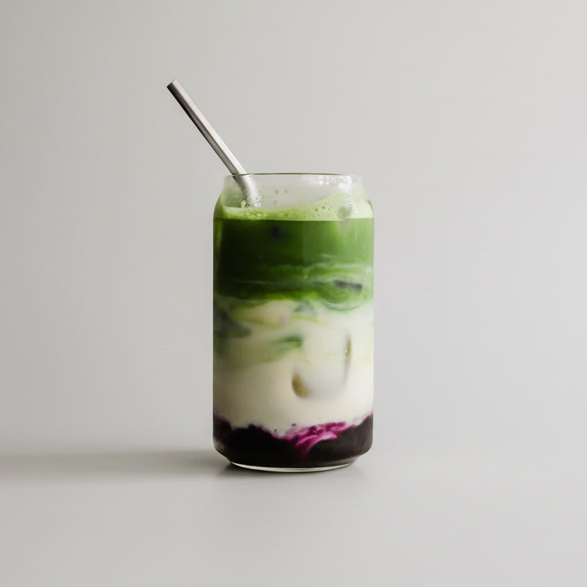 Green smoothie with a spoon in a glass, surrounded by blueberries and matcha powder on a white surface.